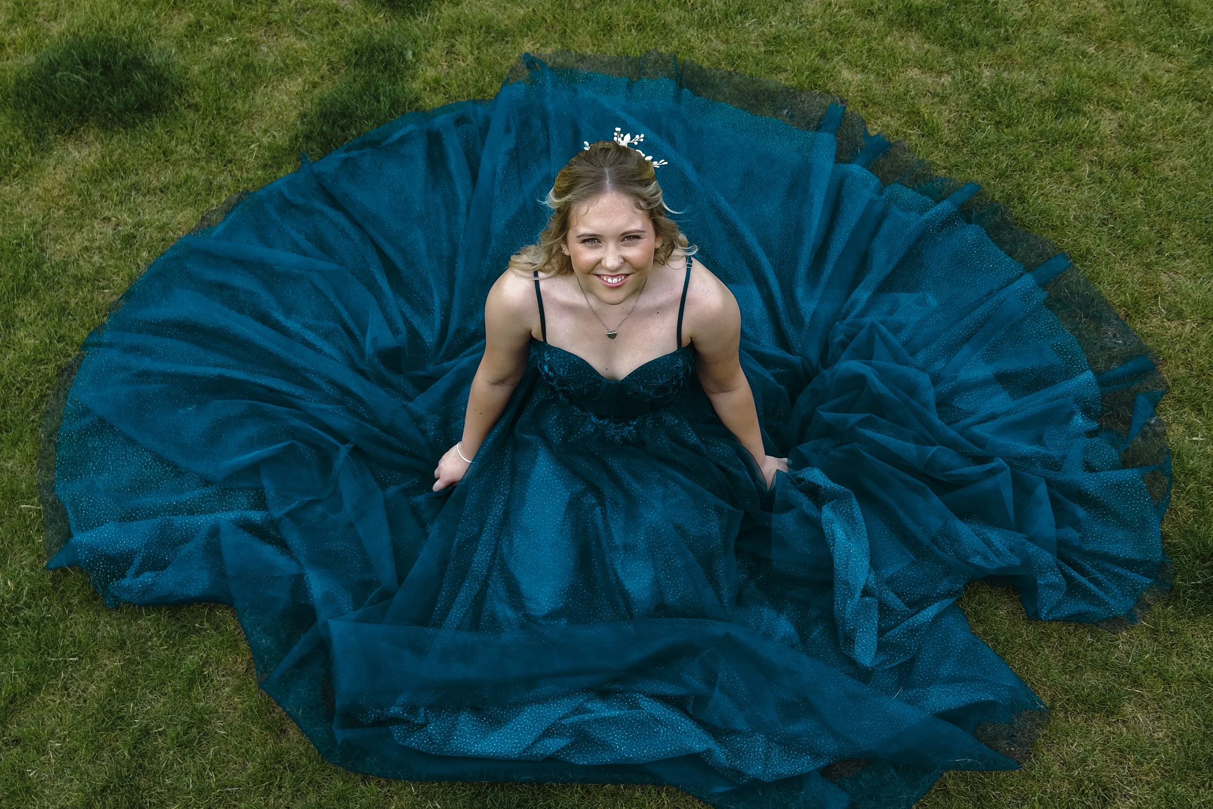 A young woman in a black dress with spaghetti straps sitting inside a circular blue tulle gown on a grassy lawn, smiling at the camera.