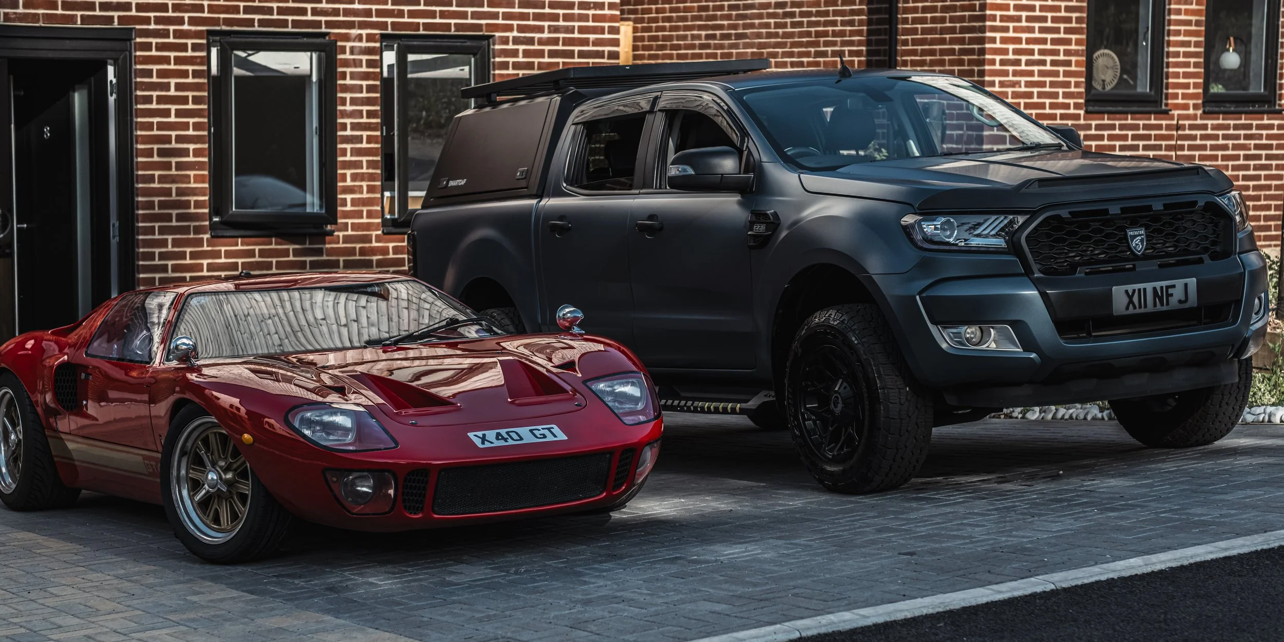 A red vintage race car with the license plate X40 GT and a black modern pickup truck with the license plate XII NFJ parked in front of a brick building with open black window frames.