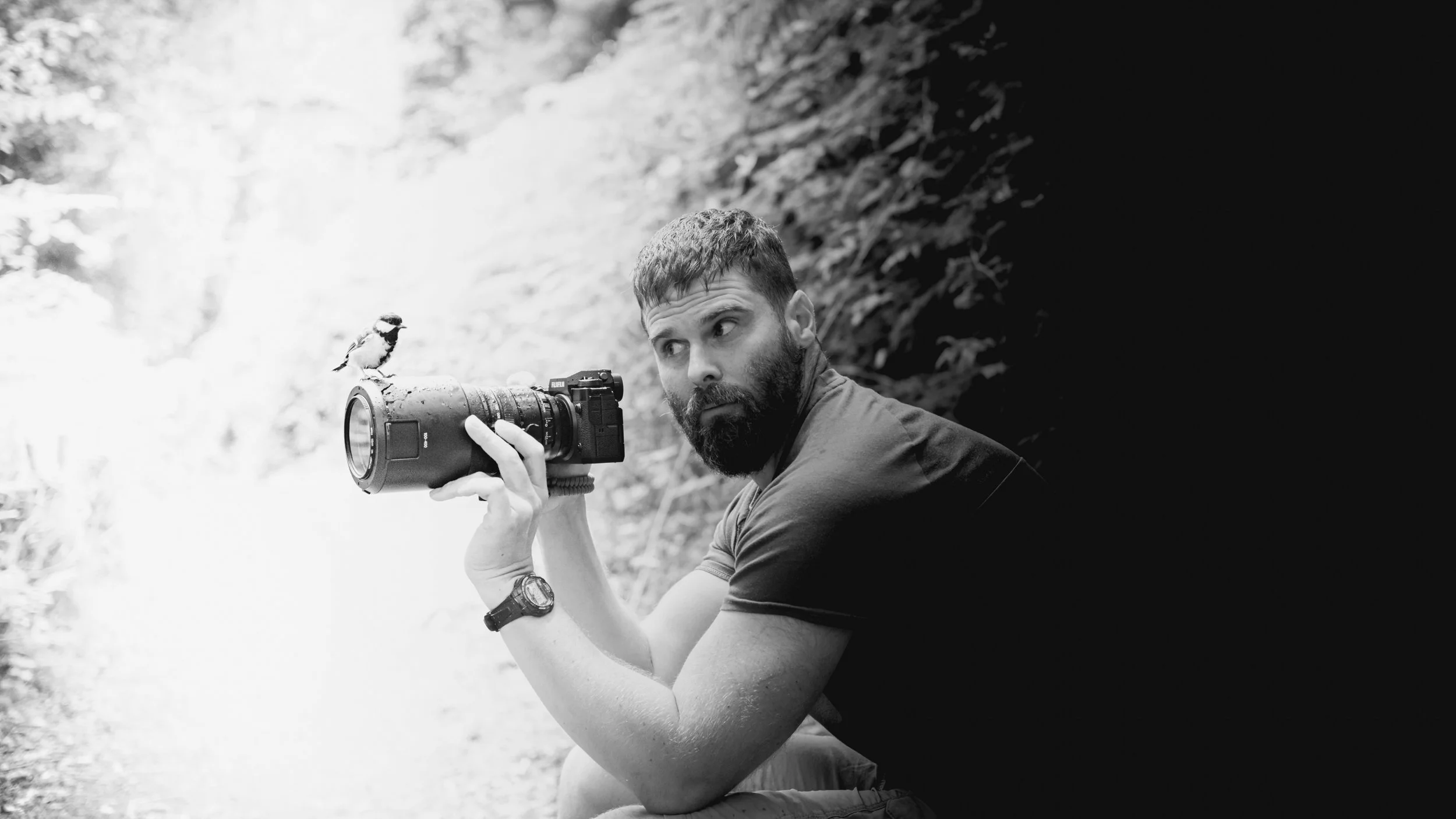 A man with a beard, holding a camera with a long lens, crouching in a forest, with a bird perched on the camera.