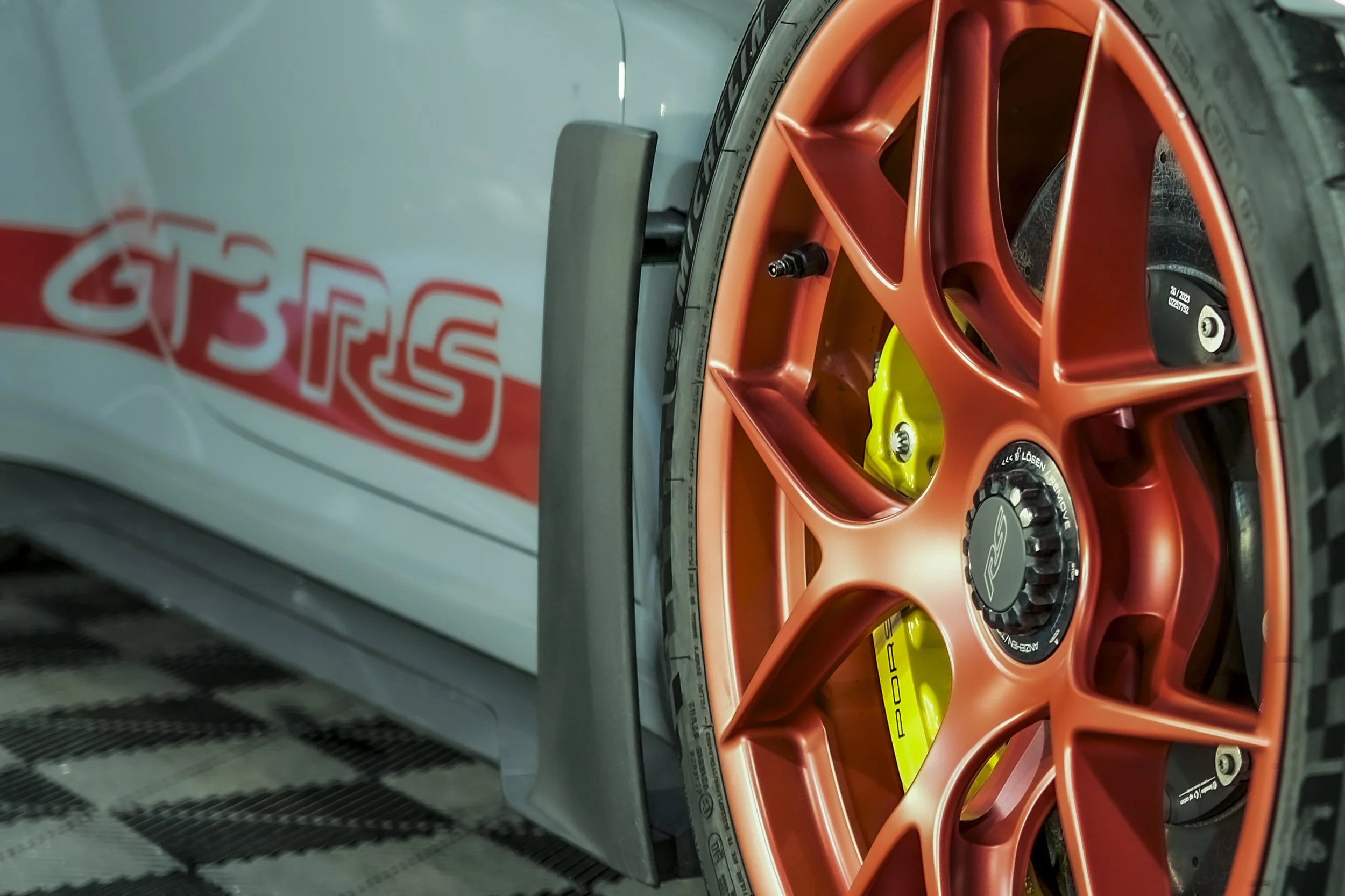 Close-up of an orange racing car wheel with a yellow brake caliper and black tire, partially blocked by a gray metallic structure with some red and white text.