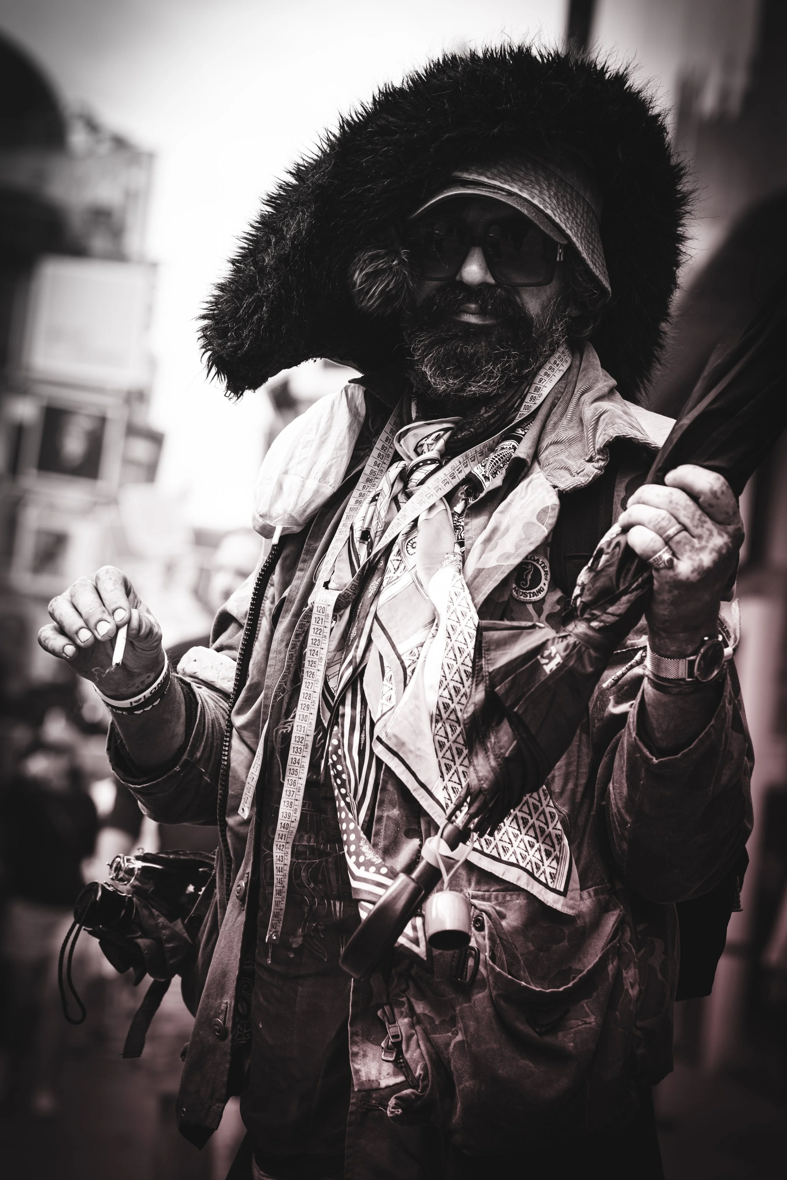 A man with a dark beard wearing a large black fur-lined hood, dark sunglasses, and layered clothing, holding objects and wearing a measuring tape around his neck, in a busy outdoor setting.