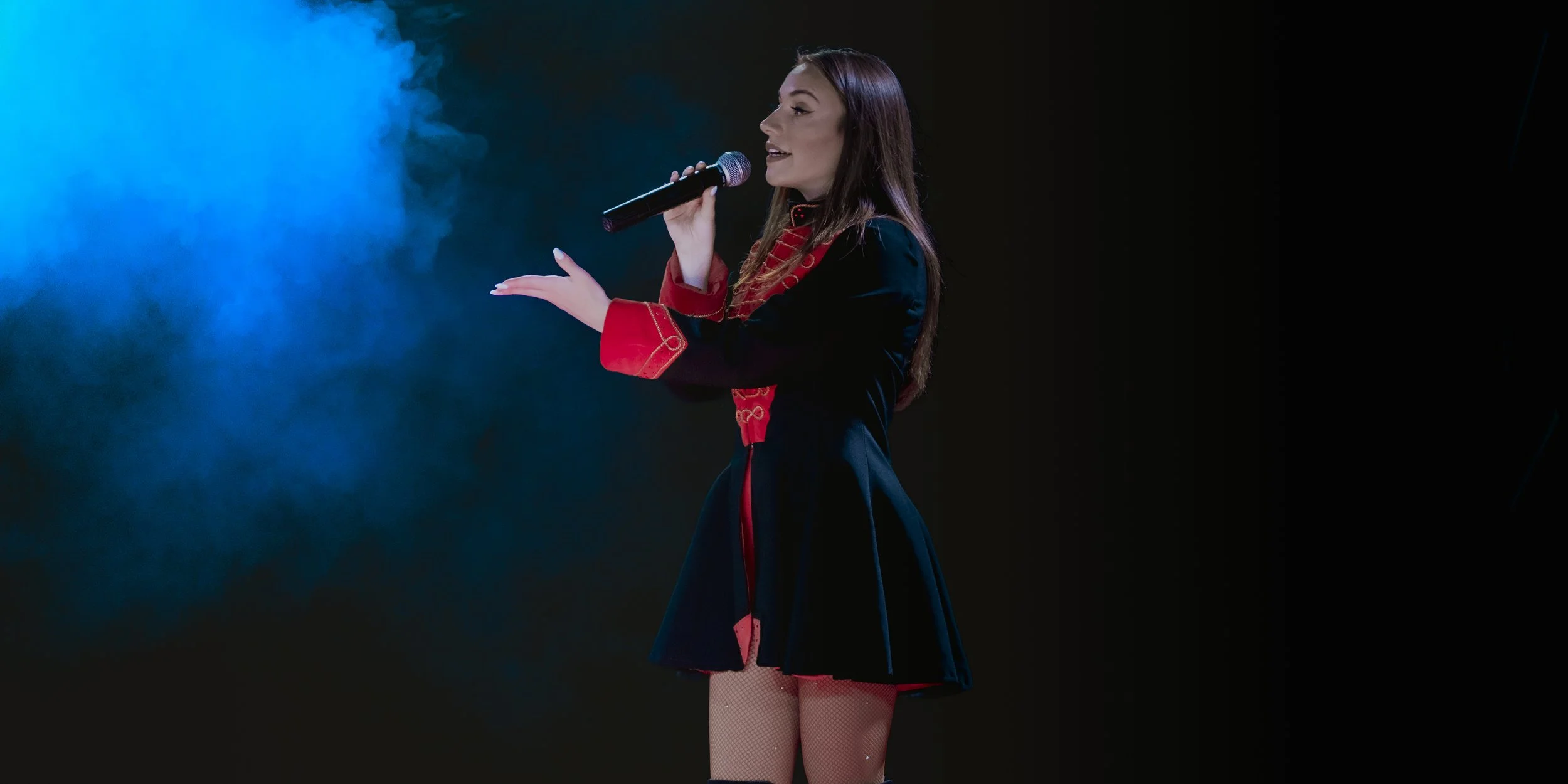 A woman with long brown hair, wearing a black dress with red accents and fishnet stockings, singing into a microphone against a dark background with blue stage fog.