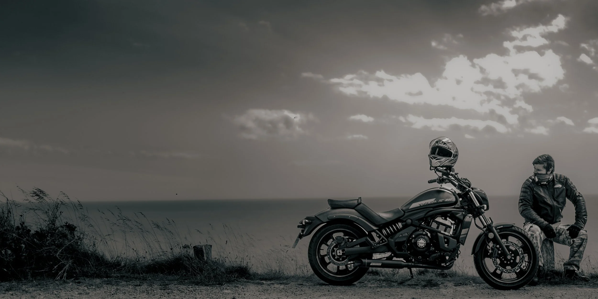 A man in motorcycle gear and a face mask sitting beside a black motorcycle on a scenic coast with cloudy sky.