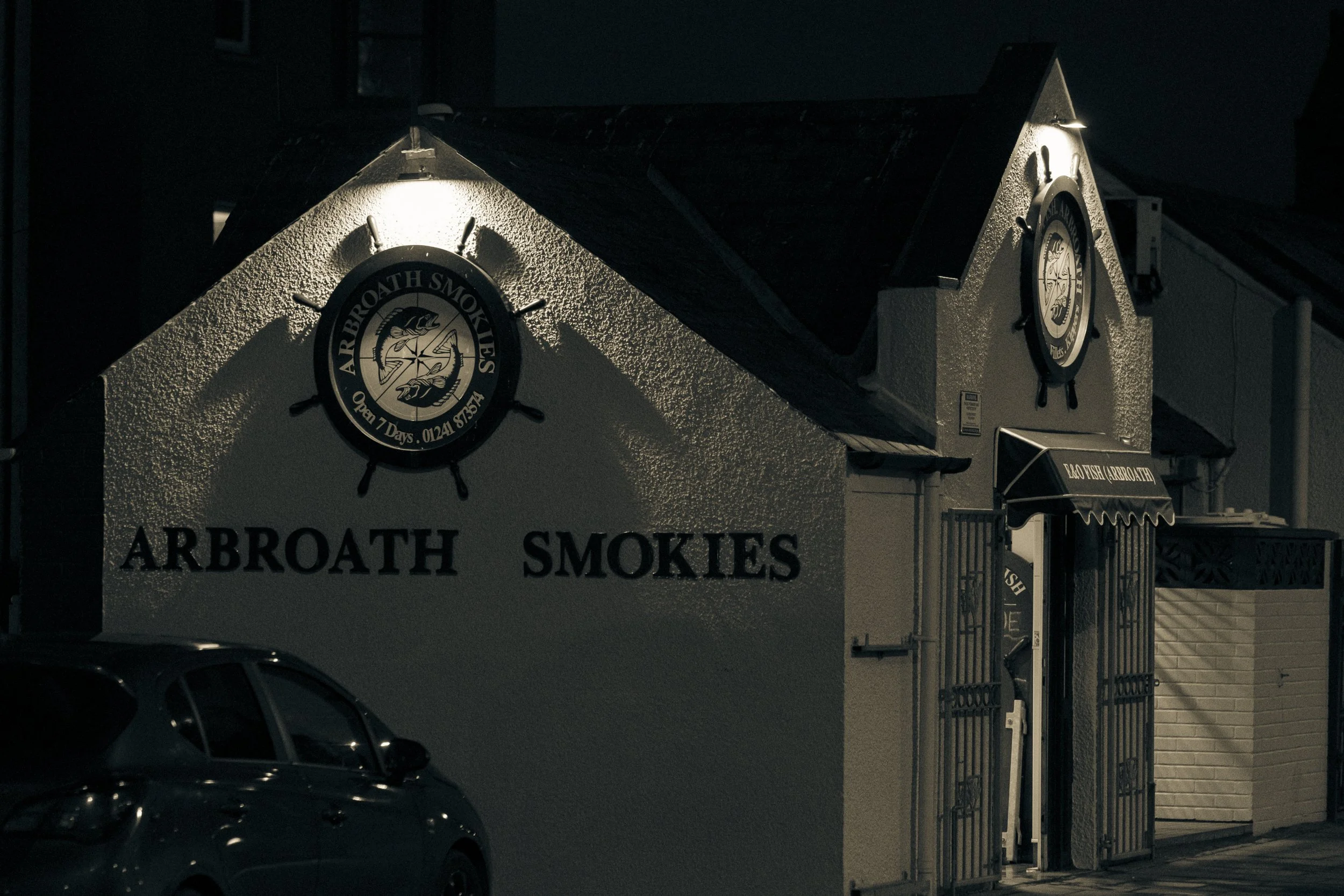 Night view of a building with signs for Arbroath Smokes and Arroath Smokehouse, illuminated by lights, with a car partially visible in front.