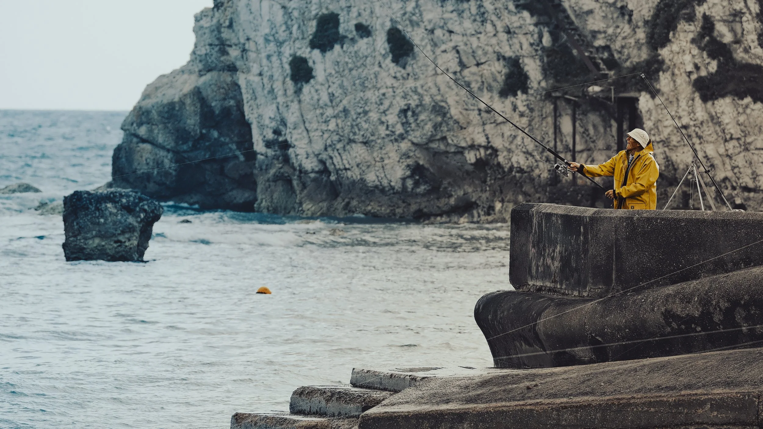 A person in a yellow jacket and white hat fishing from a concrete pier near the ocean, with a rocky cliff in the background.