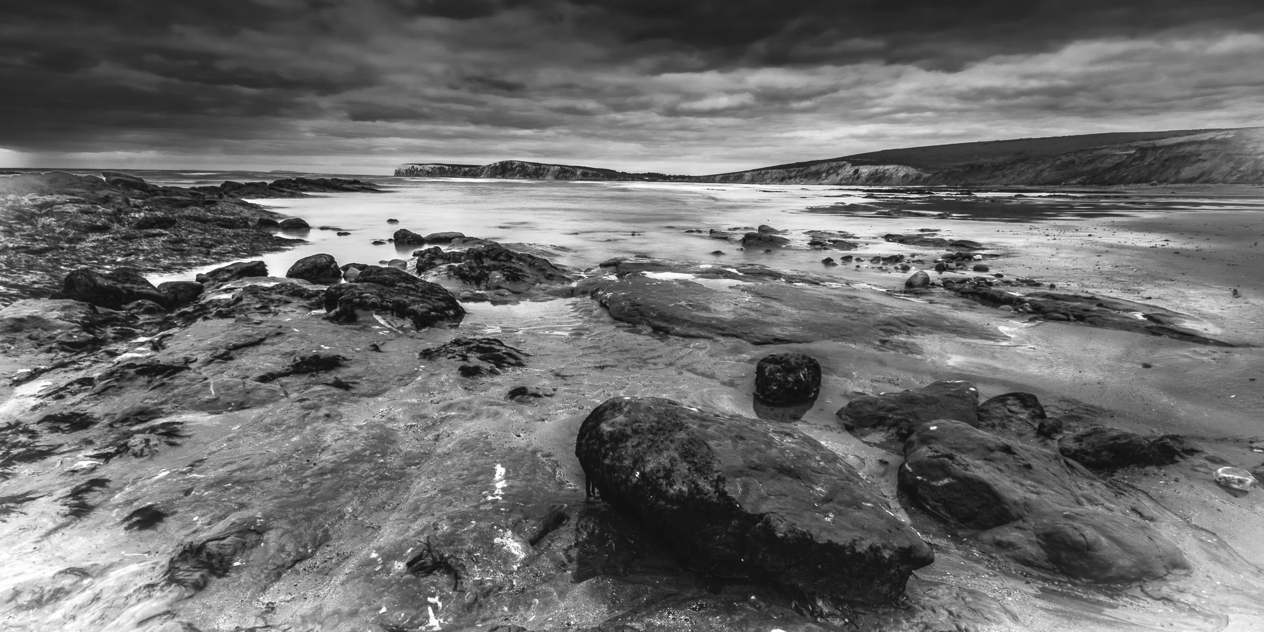 Black and white photo of a rocky beach with waves, distant cliffs, and cloudy sky.