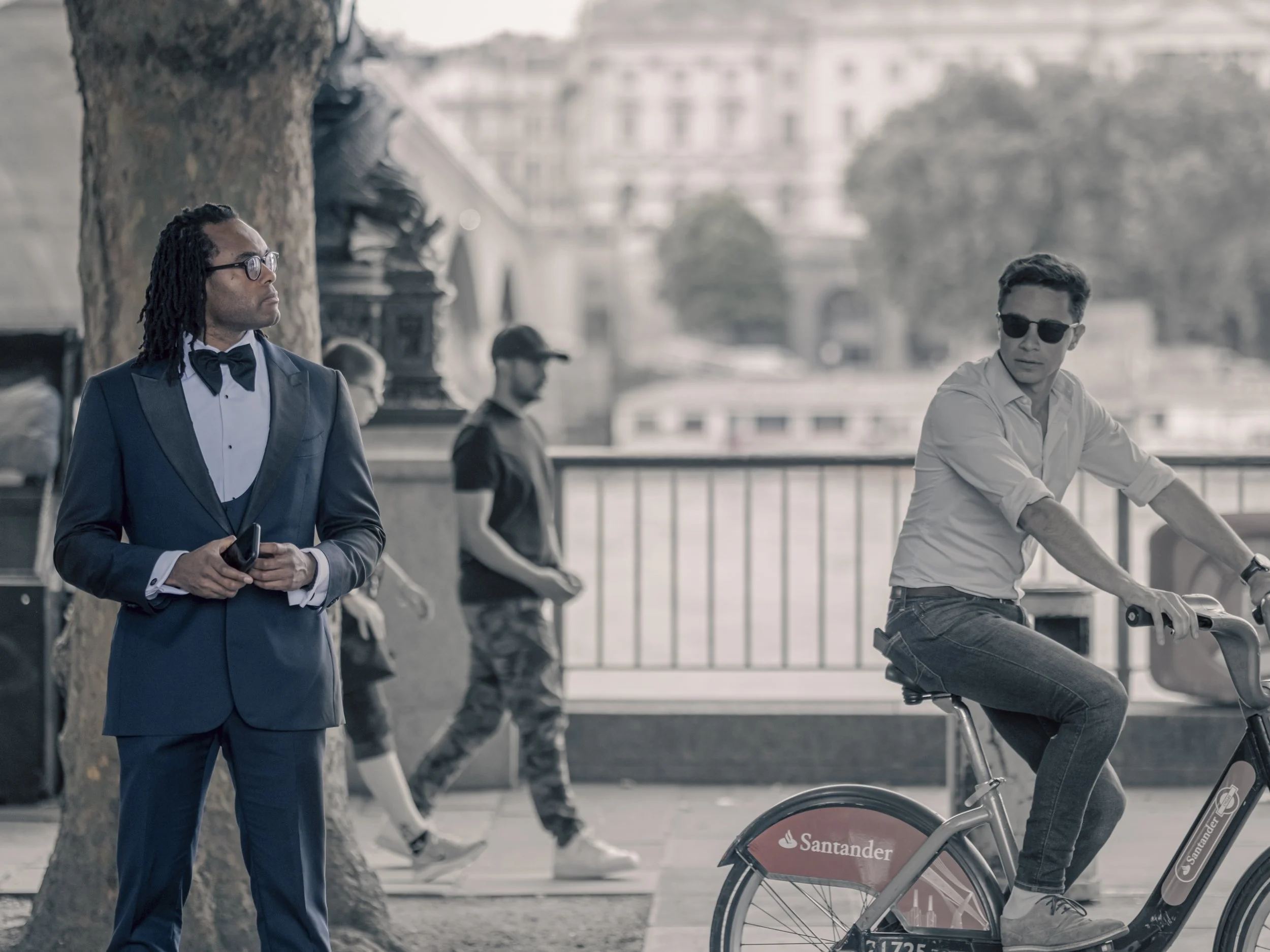 A man in a formal suit and bow tie wearing glasses stands near a tree, holding a phone, while another man in casual attire with dark sunglasses sits on a bicycle on a city street. The background shows other pedestrians and buildings.