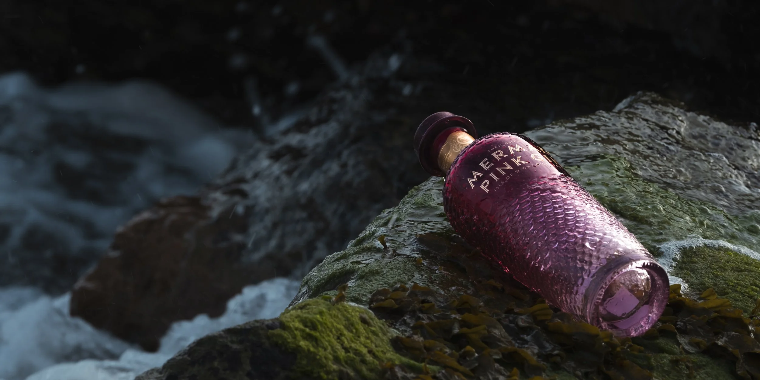 A pink textured beverage bottle labeled 'Aermark Pink' lying on rocks in a stream with flowing water.
