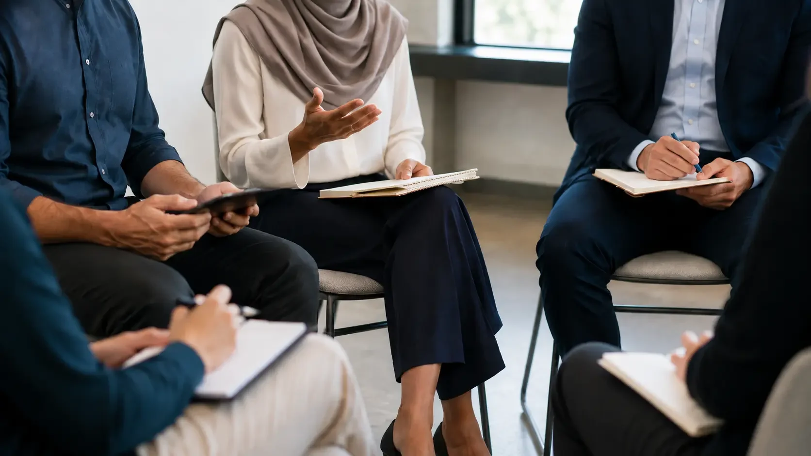 People sitting in a circle during a discussion, with notebooks and pens, in an indoor setting with natural light.