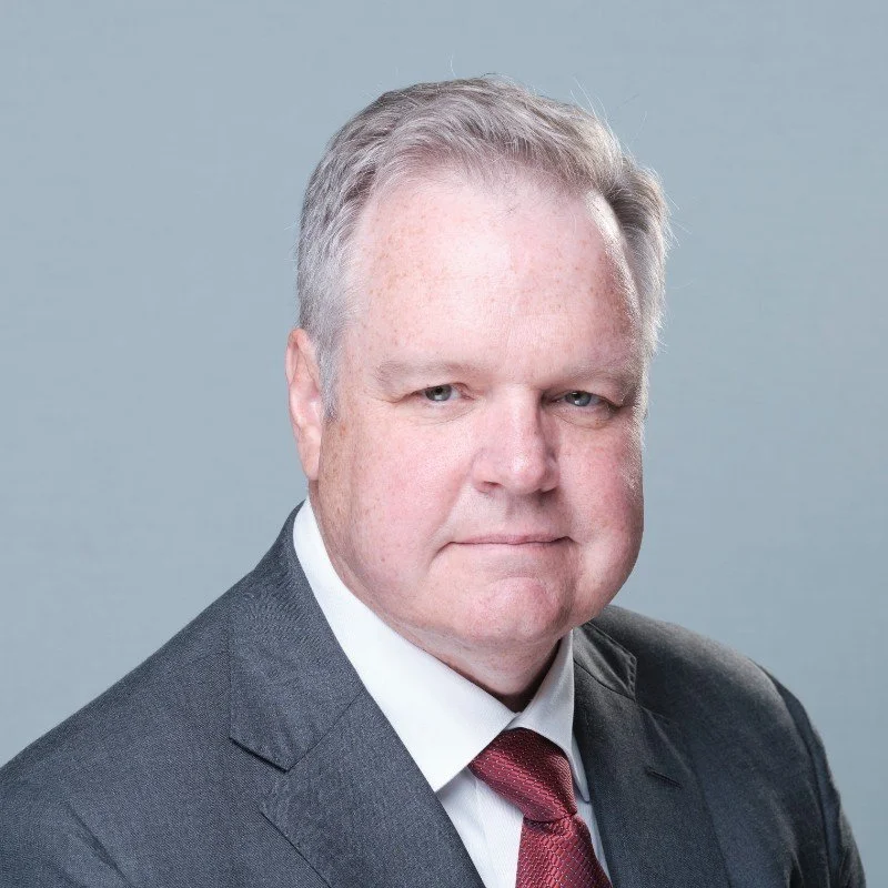 A professional headshot of a middle-aged man with light hair, wearing a dark suit, white shirt, and red tie, against a plain gray background.