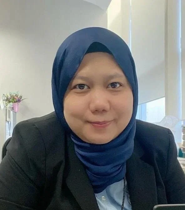 A woman wearing a blue hijab and black blazer, smiling at the camera in an office setting.