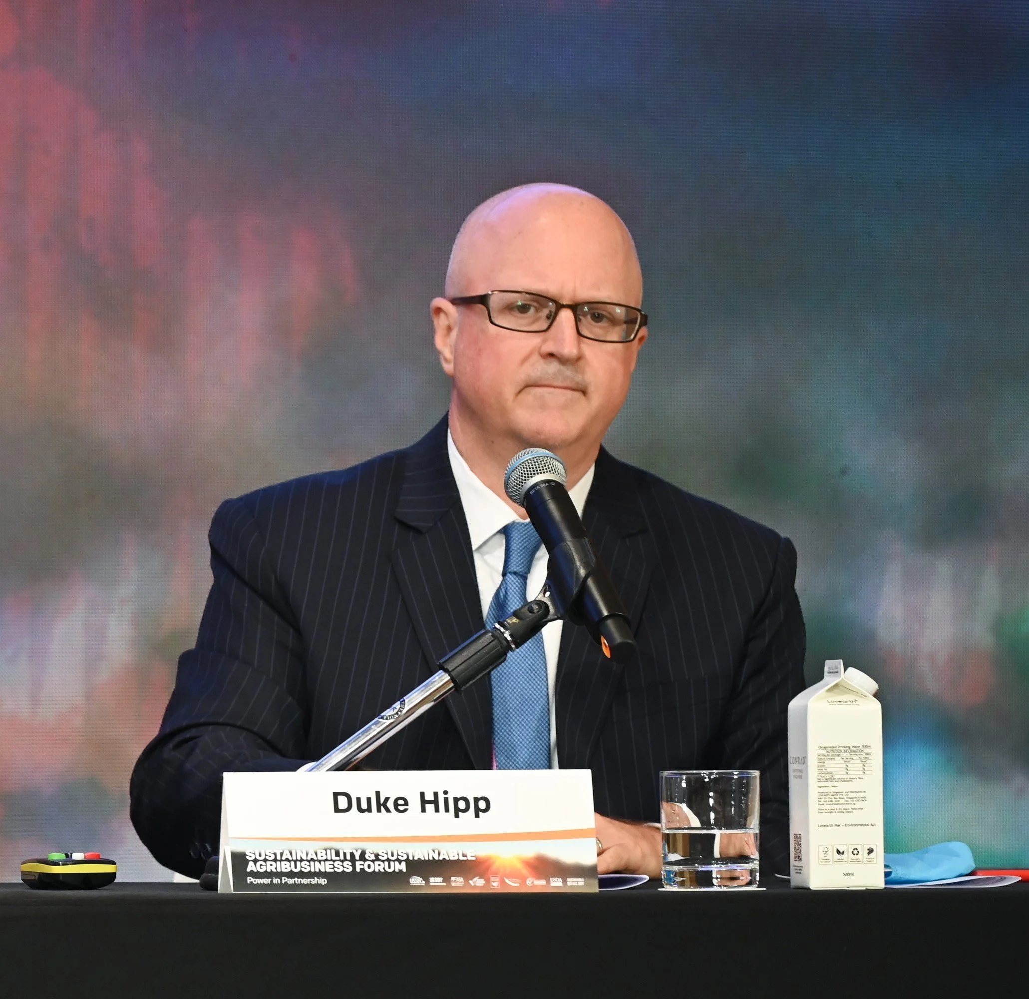 Bald man wearing glasses and a dark pinstripe suit speaking at a conference with a microphone, sitting behind a table with water bottles, a conference program, and a sign that reads Duke Hipp.