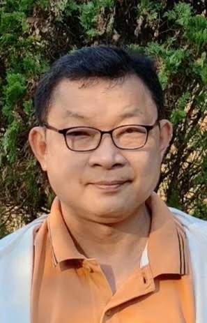 A man with short black hair and glasses, smiling outdoors with greenery in the background, wearing an orange shirt and light jacket.