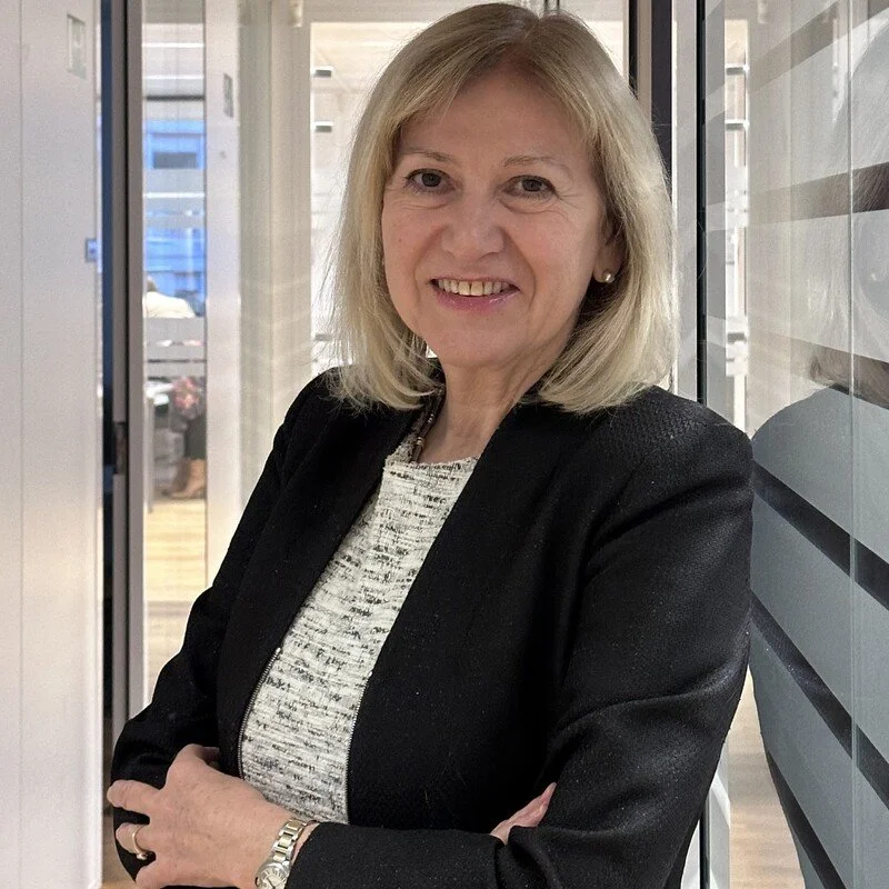 A middle-aged woman with shoulder-length blonde hair, wearing a black blazer and a light-colored top, smiling and standing with her arms crossed in a modern office corridor with glass walls.