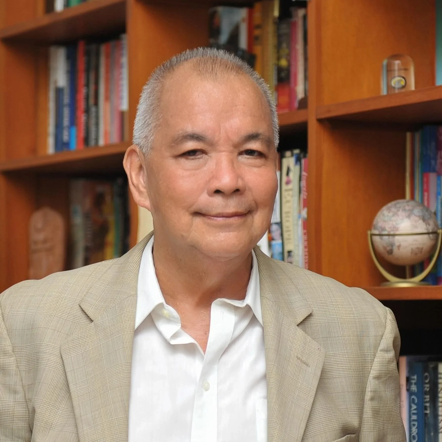 Close-up of a smiling man with short gray hair, wearing a beige checkered blazer and white shirt, in front of a bookshelf filled with books and decorative items.