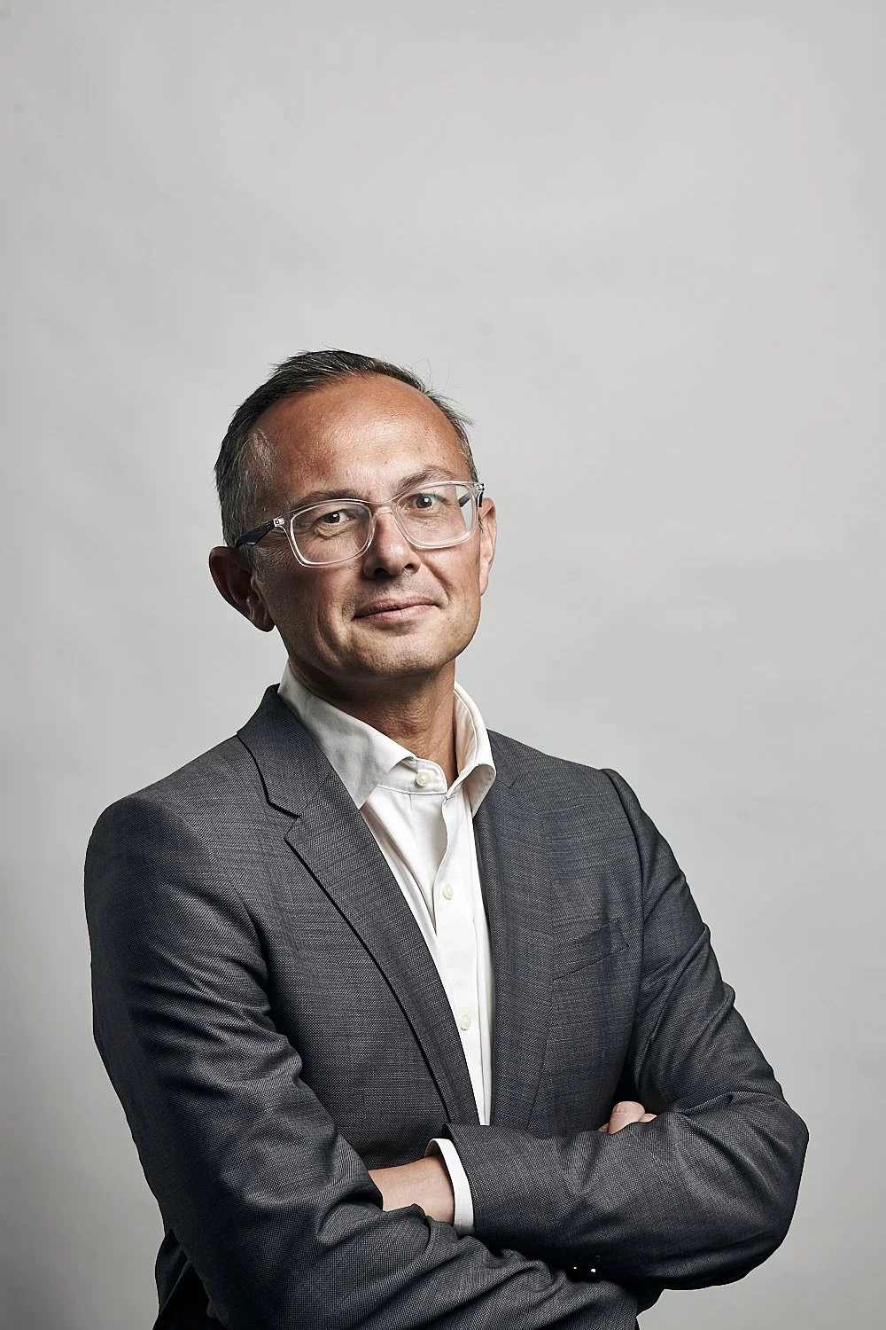 A professional man wearing glasses, a gray suit, and a white shirt, standing against a plain, light-colored background with arms crossed.