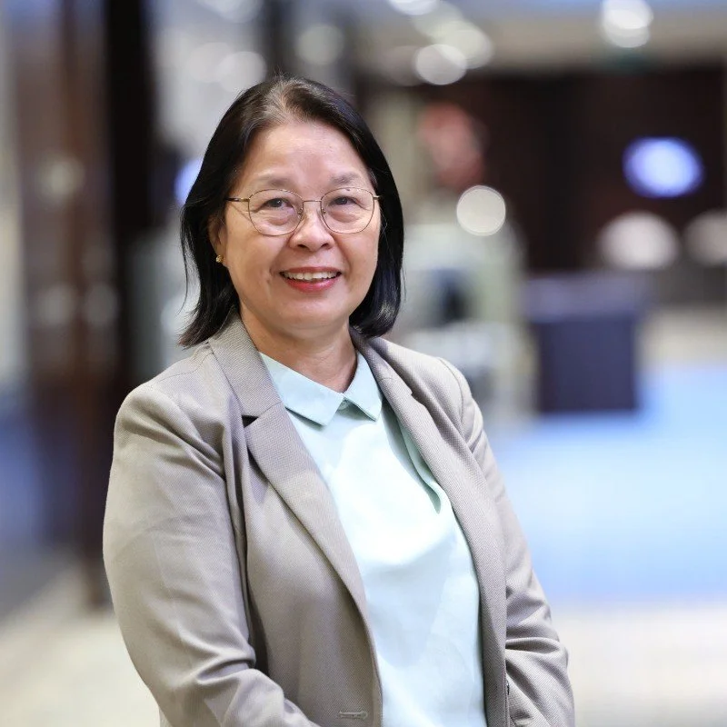 A woman with black hair and glasses smiling, wearing a blazer and light blue shirt, standing indoors with a blurred background.