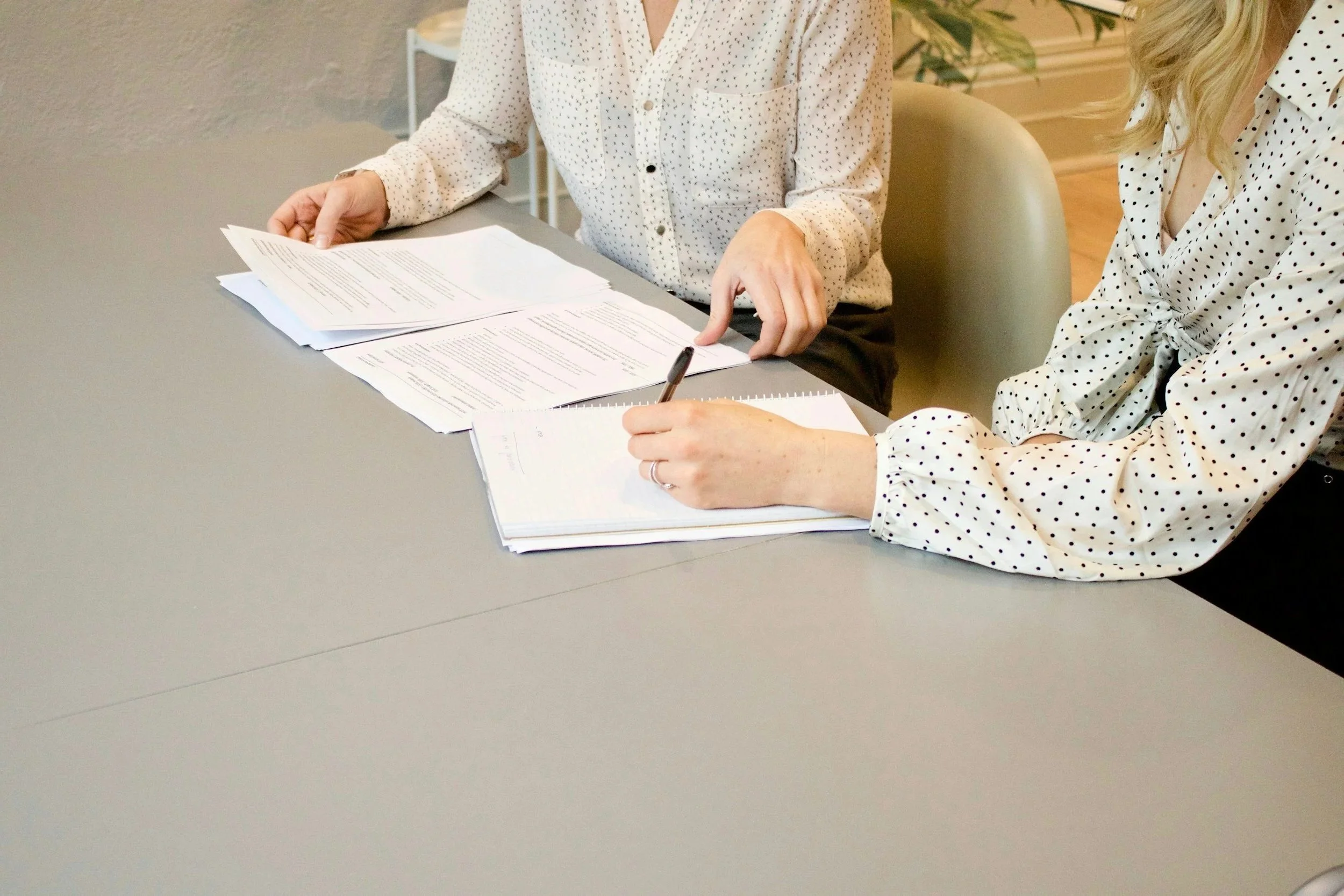 Two women sit at a table reviewing and taking notes on paperwork and documents.