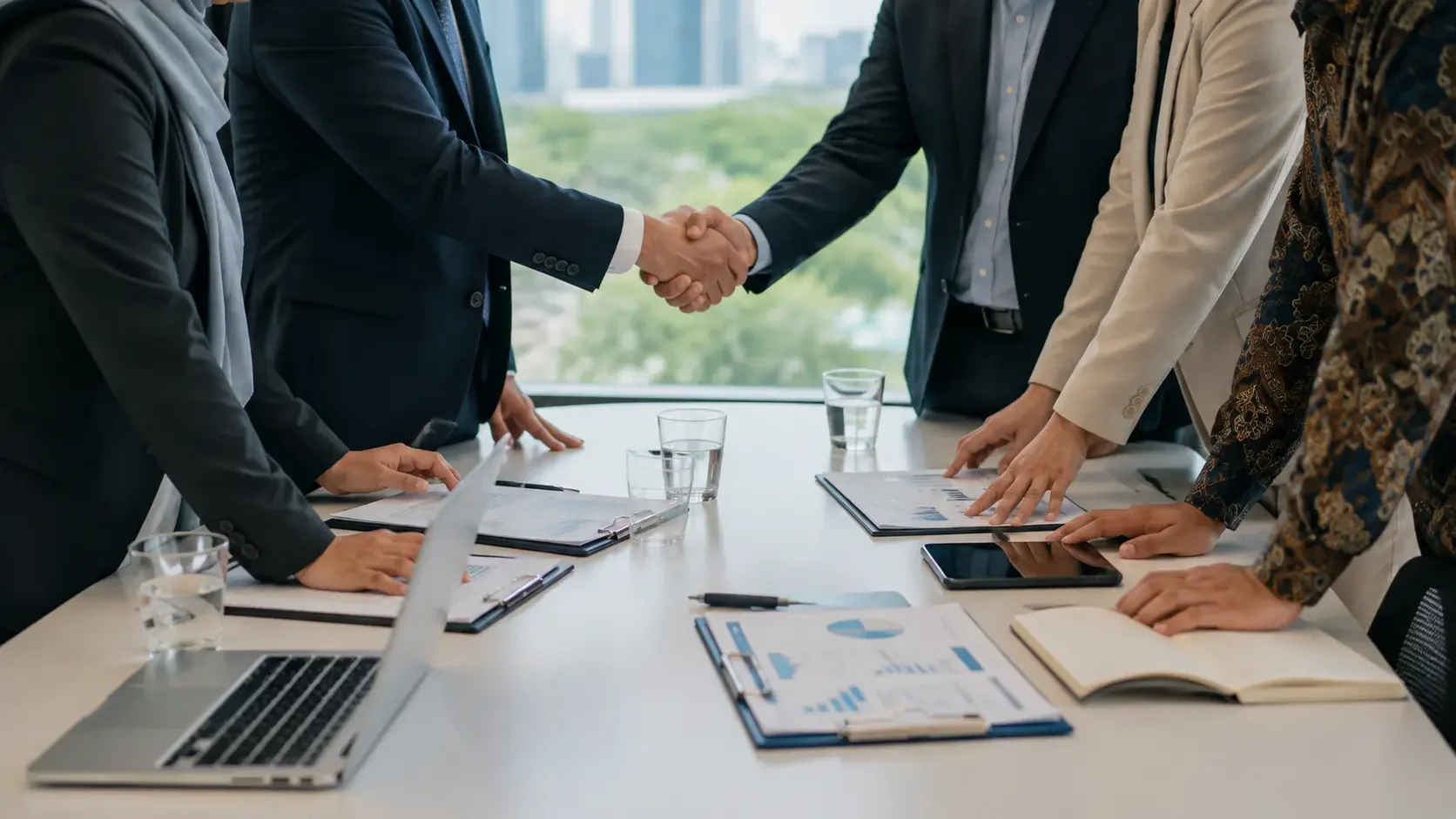 Businesspeople shaking hands over a conference table with laptops, documents, tablets, and glasses of water, in a modern office with large windows.