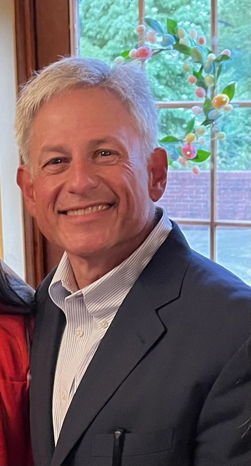 Smiling man with gray hair in a suit, standing in front of a window with a decorative hanging ornament of colorful round beads and green leaves.