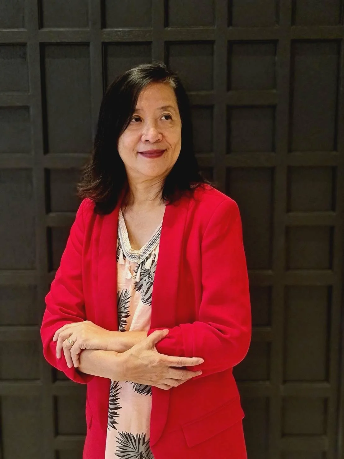 A woman with shoulder-length black hair wearing a red blazer and a patterned blouse, standing against a dark grid-patterned wall.