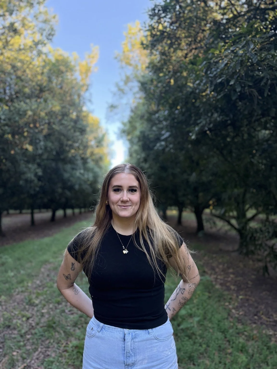A young woman with long hair and tattoos on her arms standing outdoors in a park with trees and a pathway, smiling at the camera.