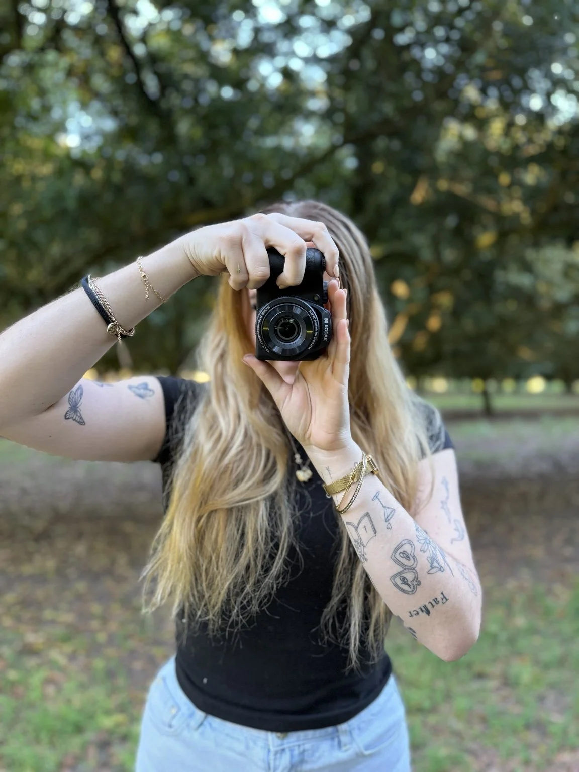 A person with long wavy blonde hair taking a photo outdoors with a camera, standing in a park with green trees in the background.