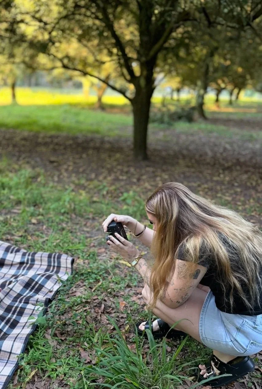 A woman with long, wavy hair crouches on the grass, holding a camera and taking a photo in a park with green trees and grass.