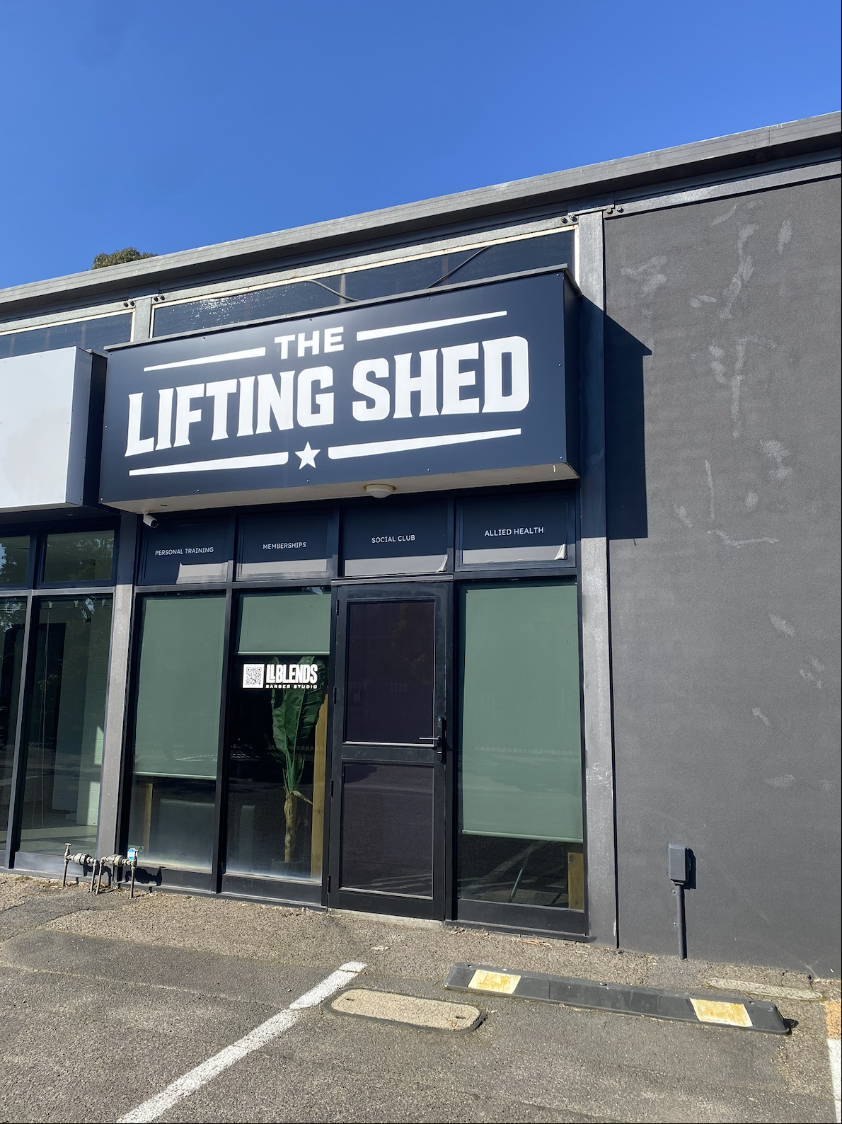 Front view of a building with a sign that reads 'The Lifting Shed'. The building has large windows with smaller signs above, including 'Personal Training,' 'Memberships,' 'Social Club,' and 'Allied Health'. There is a QR code and sign 'LBLENDS BARBER STUDIO' on the door, and a small garden with a plant inside.