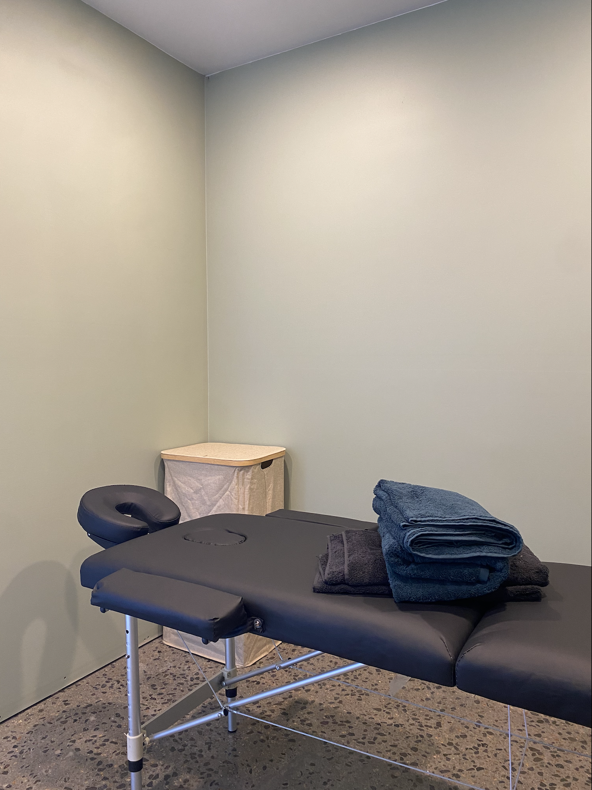 Massage therapy room with a black massage table covered in folded blue and dark gray towels, a rolled-up towel on top, a small beige storage bin in the corner, and mint green walls.