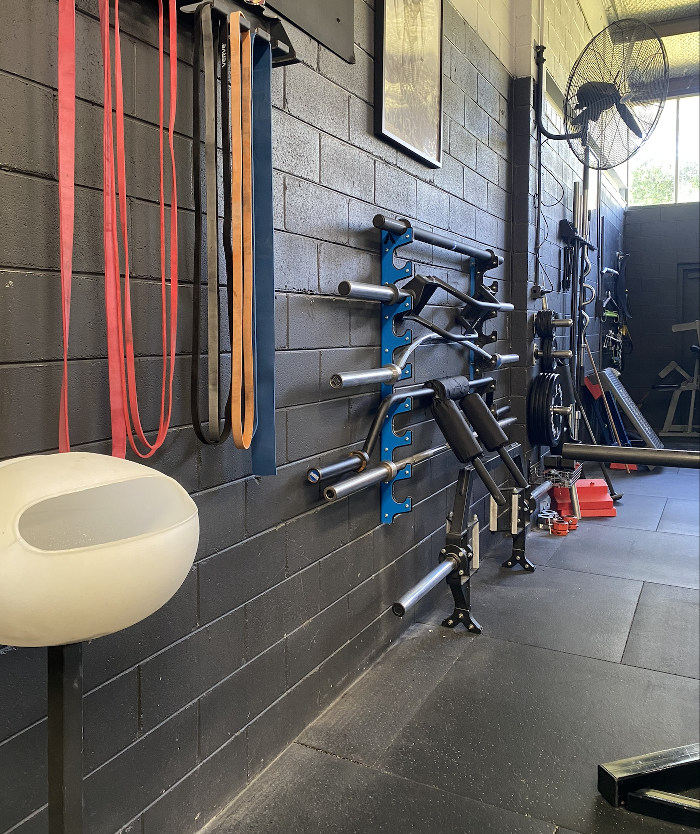 Weightlifting gym with various exercise equipment including resistance bands, barbells, weights, and a fan on a brick wall.
