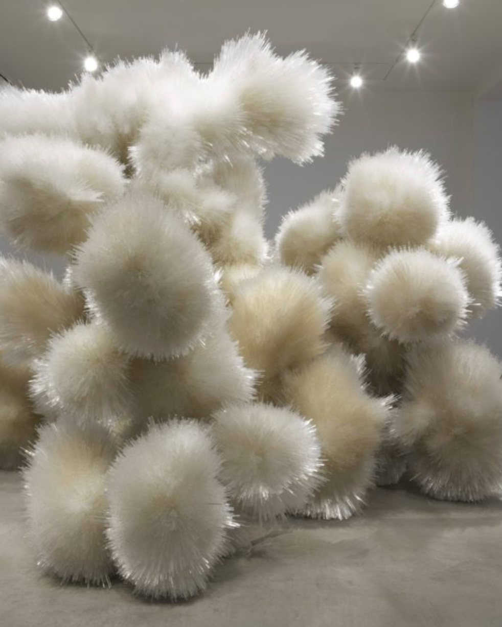 A collection of white fluffy, spherical sculptures with spiky textures, displayed in an art gallery with ceiling lights.