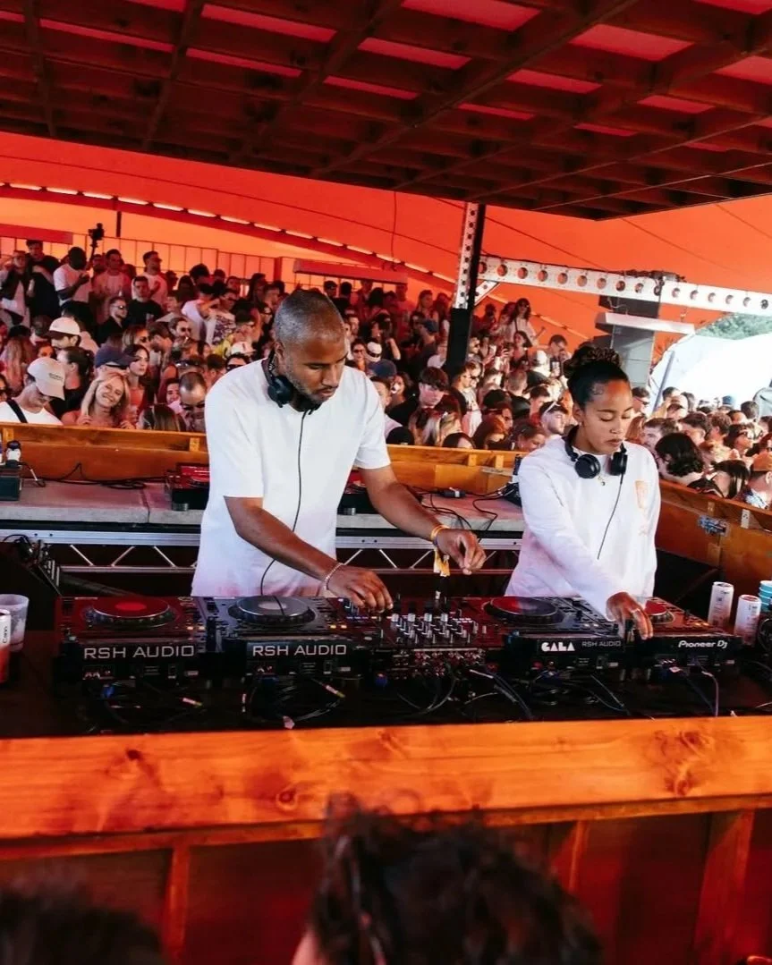Two DJ performers are mixing music on a DJ console at an outdoor event with a large audience under an orange canopy.