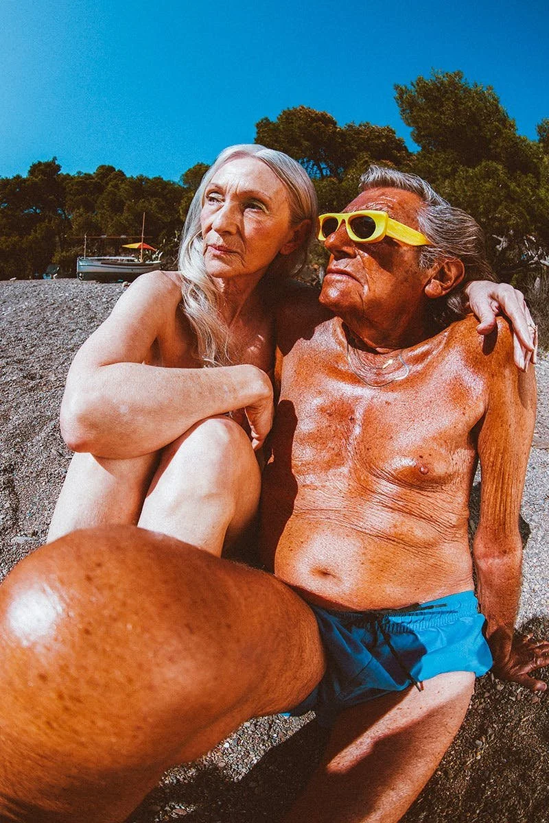 Two elderly people sitting on a rocky shore, one wearing sunglasses and the other with long gray hair, with a boat and trees in the background on a sunny day.