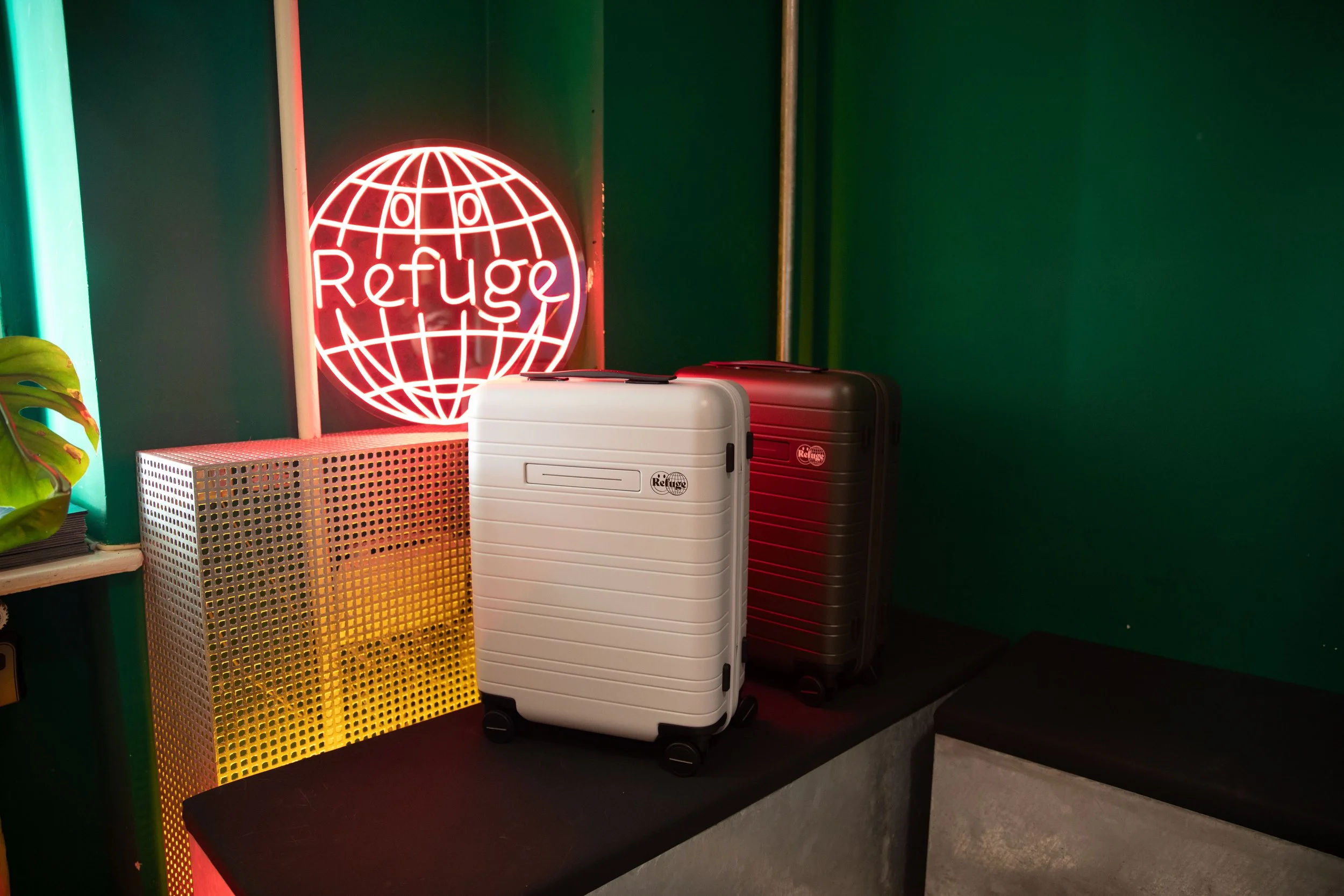 Neon sign with a globe and the word "Refuge" on a green wall, with two suitcases and a yellow mesh container on a black table.