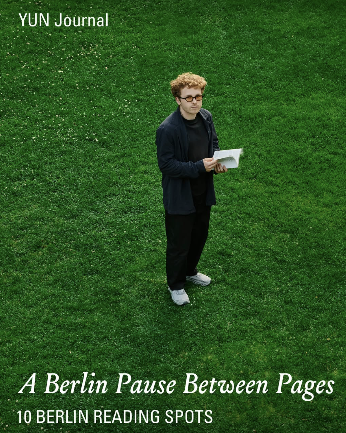 Cover of YUN Journal titled "A Berlin Pause Between Pages" featuring a man with curly hair and glasses, wearing a dark jacket and black pants, holding an open book, standing on lush green grass.