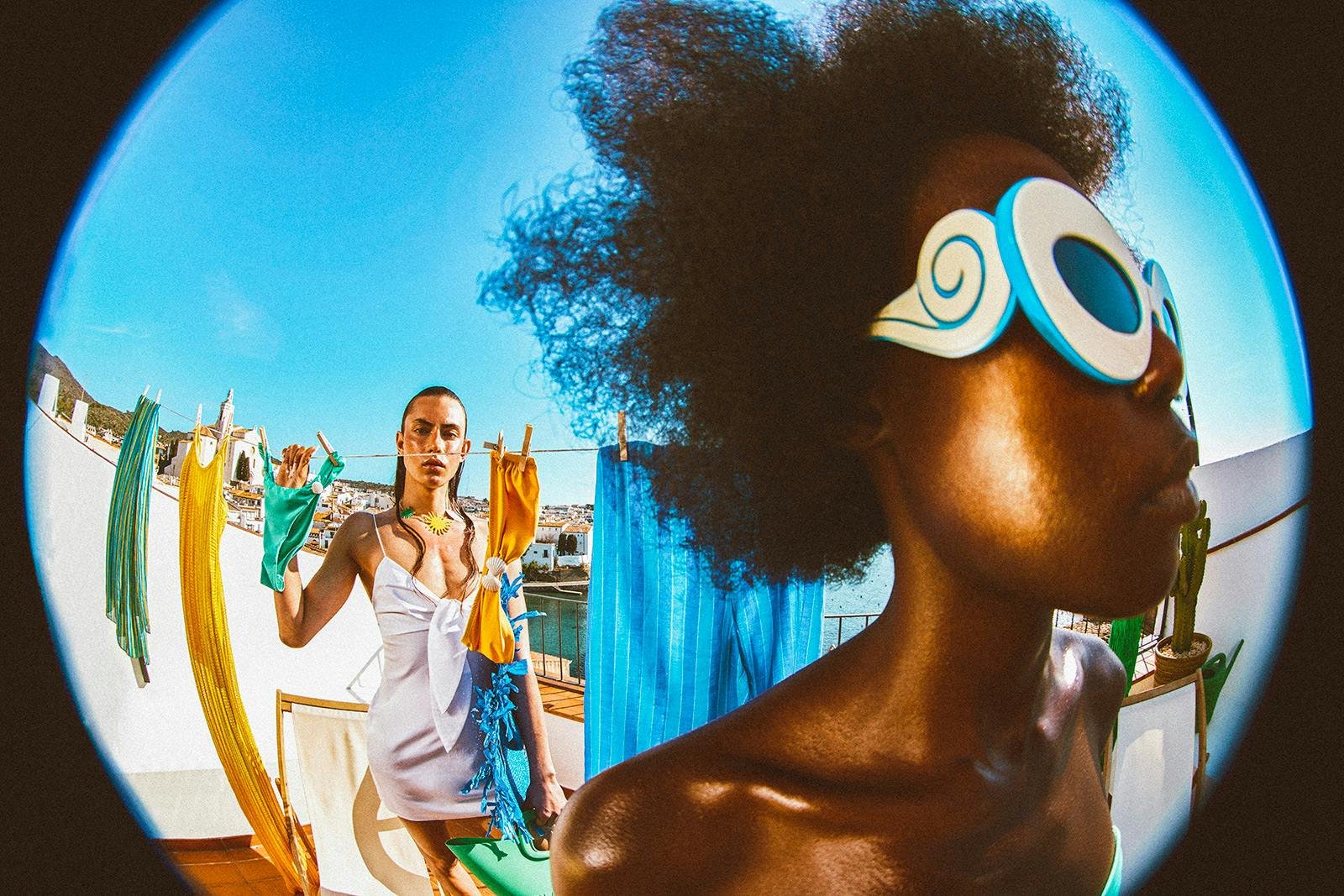 A woman with large curly hair and sunglasses stands in the foreground, with colorful laundry hanging on a clothesline behind her. The photo is taken with a fisheye lens, creating a curved perspective on a rooftop with a city and water view in the background.