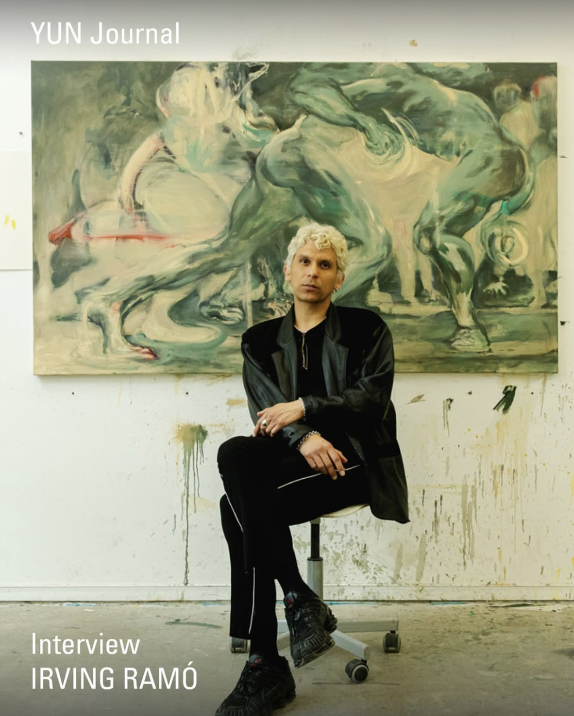A person with blond curly hair sitting on a stool in front of an abstract painting with horse figures. The person is wearing a black jacket and black shoes. The background wall has paint splatters and streaks.