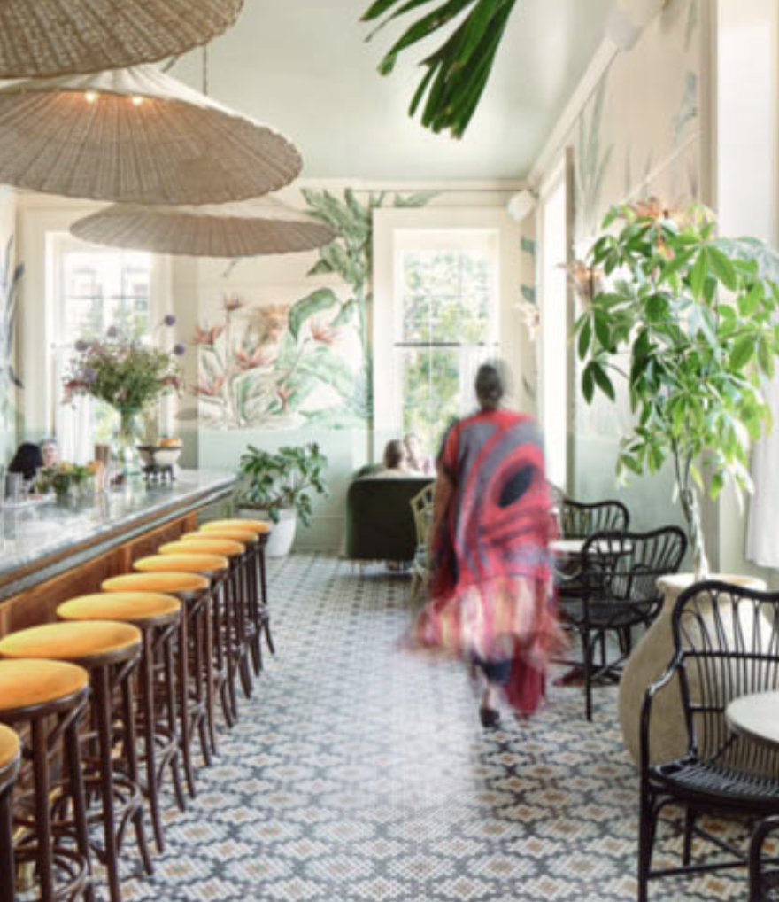 Interior of a brightly lit cafe or restaurant with a bar on the left side, a wall mural with floral and tropical designs, and various green plants. A woman in a red and black dress walks away from the camera towards other patrons near large windows.