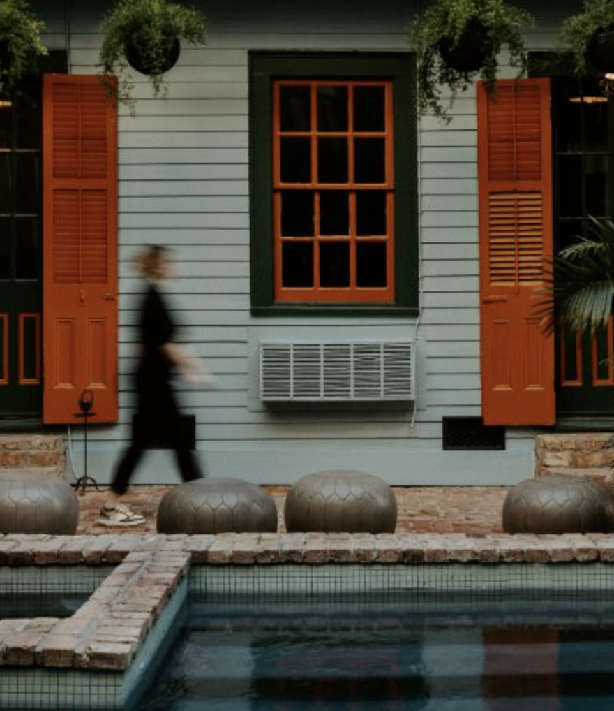 A person walking past a house with blue siding, orange shutters, and a large window with orange framing. There is a window air conditioning unit beneath the window. In front of the house, there are large rounded decorative objects and a brick-paved area with a small pool.
