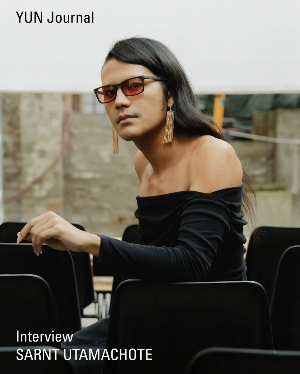 A person with long black hair wearing orange-tinted glasses and large tassel earrings, dressed in a black off-shoulder top, sitting on black chairs outdoors with a background of a stone wall and some trees. The image is titled 'YUN Journal' and marked as an interview with Sarnt Utamachote.
