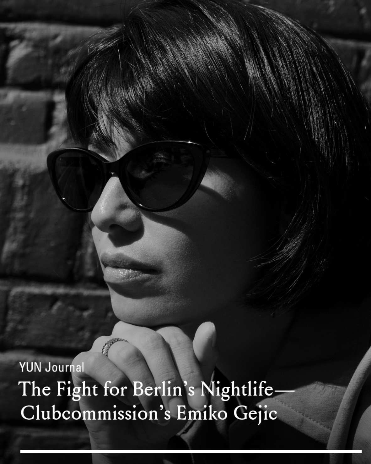 A black and white photo of a woman with short dark hair wearing sunglasses. She is resting her chin on her hand, with her fingers near her lips. The background appears to be a brick wall. The text at the bottom reads, "YUN Journal The Fight for Berlin's Nightlife—Clubcommission's Emiko Gejic."