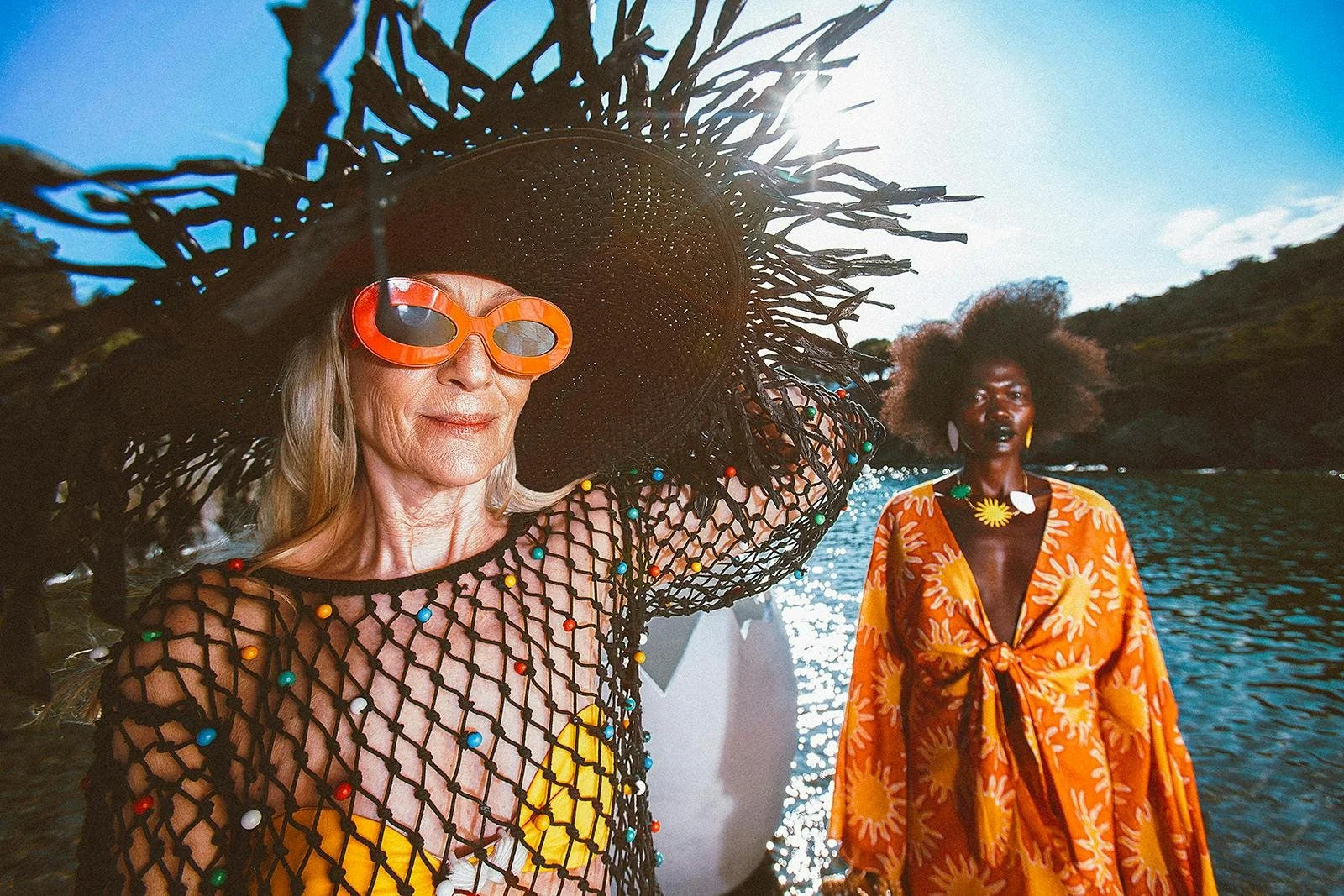 Two women dressed in colorful, bold fashion standing by a body of water with trees and a blue sky in the background. The woman on the left has blonde hair, large orange sunglasses, a broad-brimmed black hat, and a fishnet cover-up adorned with colorful beads over a yellow top. The woman on the right has dark skin, voluminous natural hair, and wears a bright orange dress with yellow and white floral patterns, accessorized with large earrings and bold jewelry.