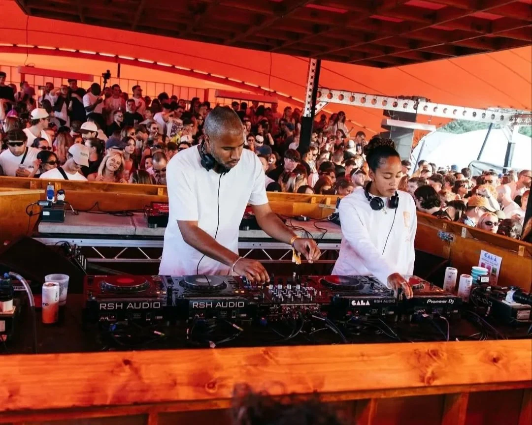 Two DJs performing at an outdoor music event, with a large crowd of people under an orange canopy.