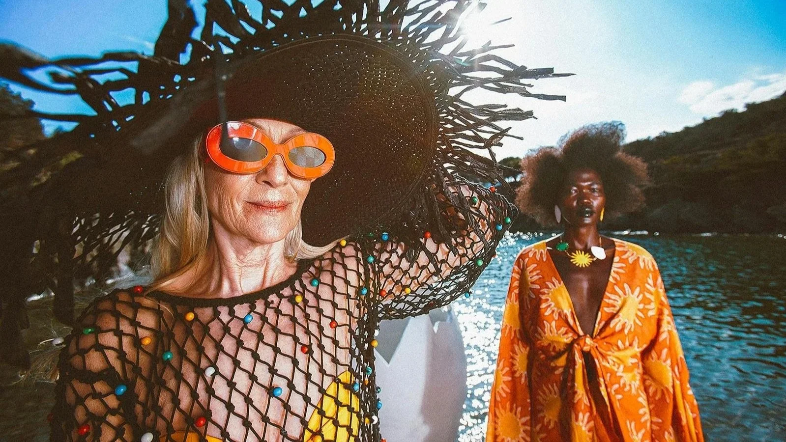 Two women standing outdoors near a body of water with hills in the background during daytime. The woman in the foreground is wearing a large floppy hat, orange sunglasses, a black fishnet top with colorful beads, and a yellow outfit underneath. The woman in the background has dark skin, an afro hairstyle, a bright orange dress with a sun pattern, and a yellow necklace and earrings.