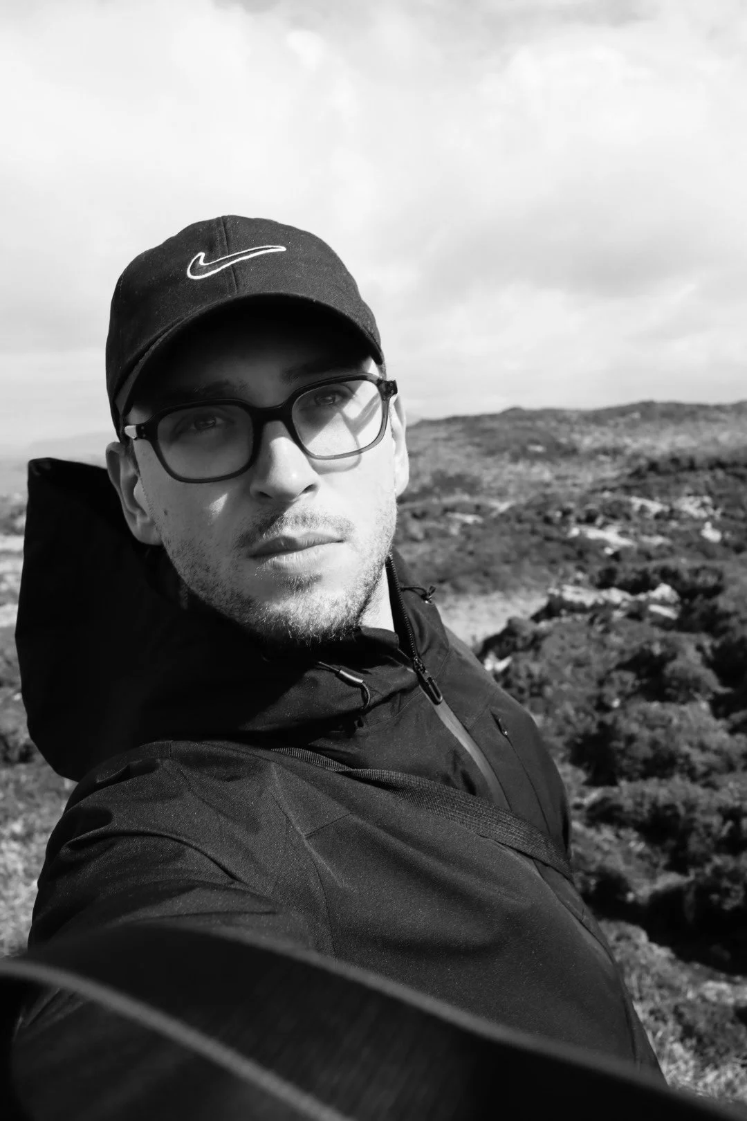 Black and white photo of a young man wearing glasses, a Nike cap, and a jacket, outdoors in a hilly landscape with cloudy sky.