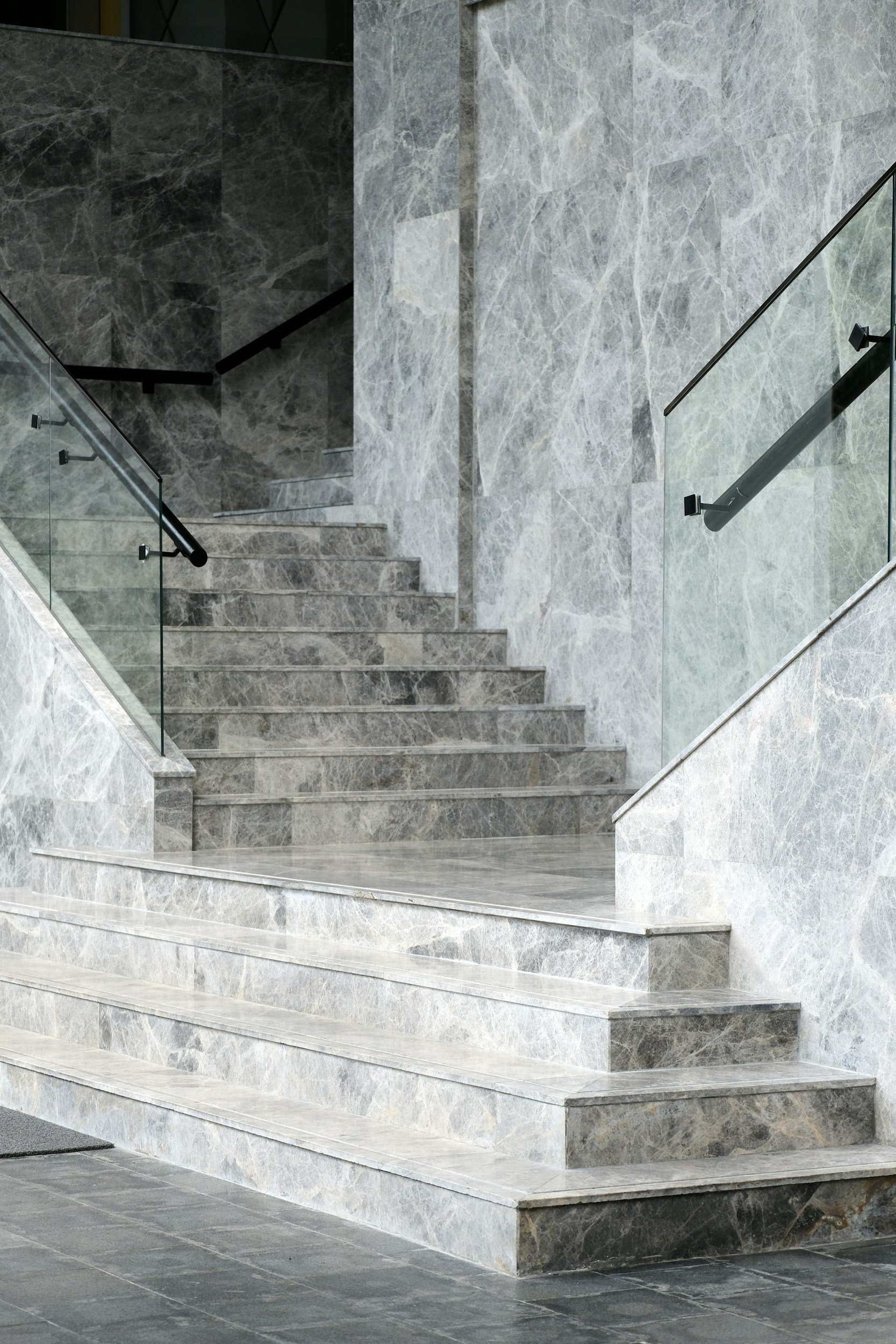 A staircase made of gray marble with a black handrail and glass panels on the sides. The wall adjacent to the staircase is also covered in gray marble with white veining.