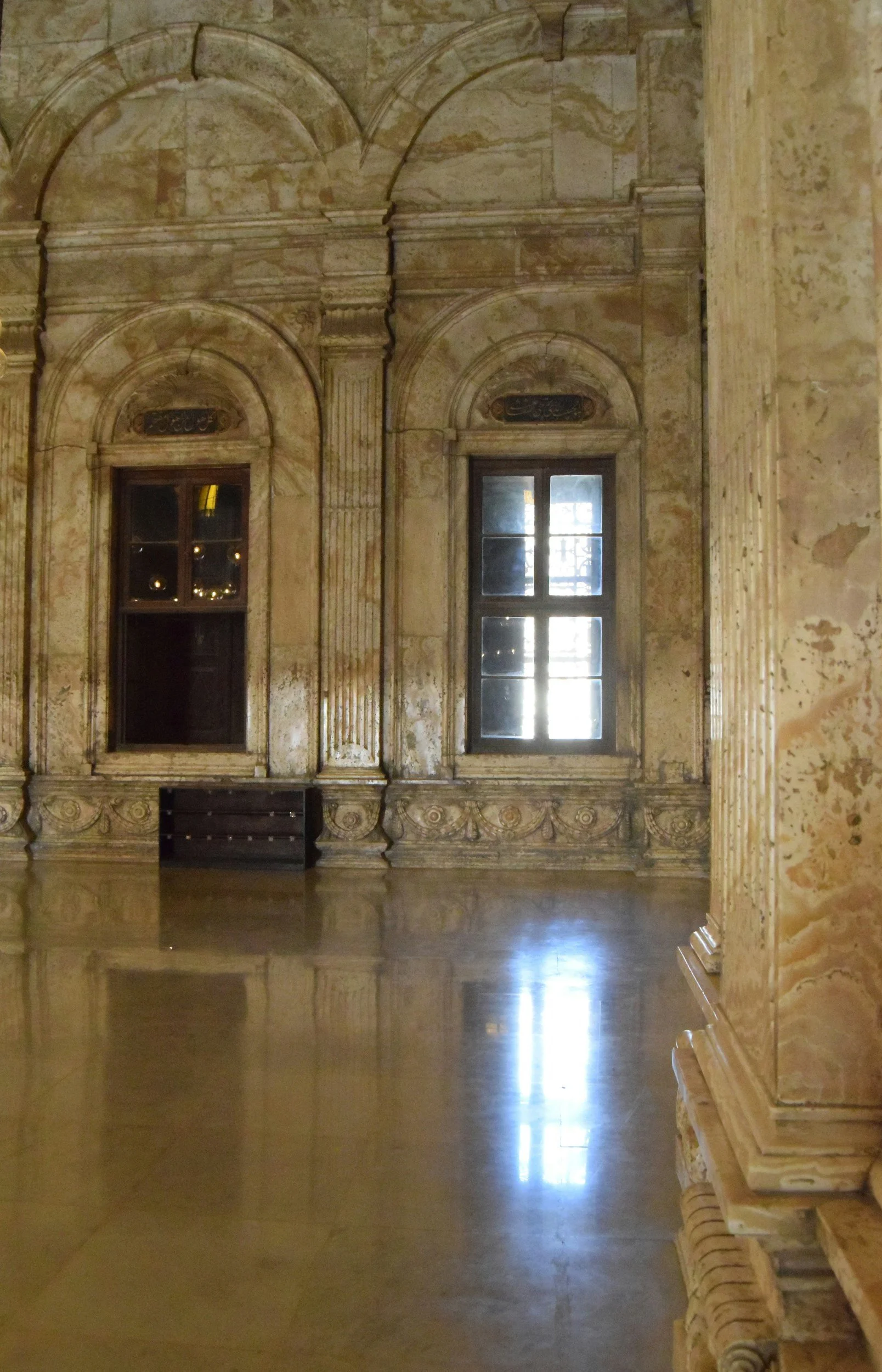 An indoor room with beige and brown marble walls and decorative architectural details, including tall windows with wooden frames, lace curtains, and reflections on the polished marble floor.