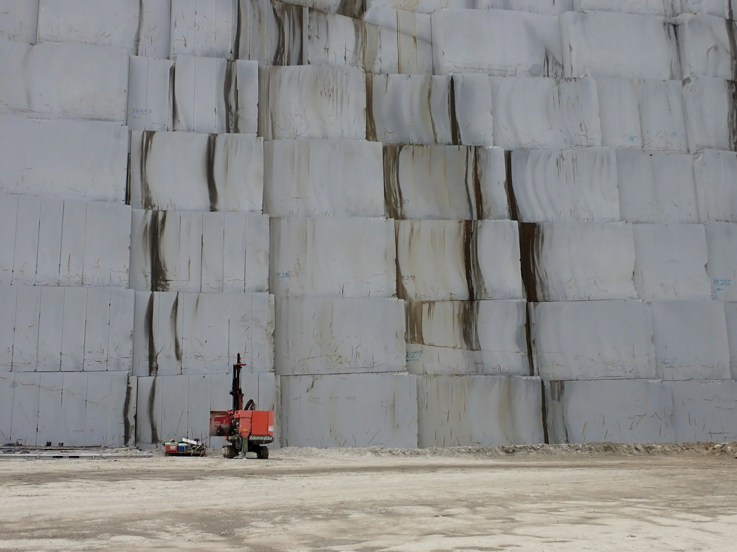A large white marble quarry wall with visible veins, and construction equipment, including a small red crane and concrete mixer, on a dirt ground in front of the wall.