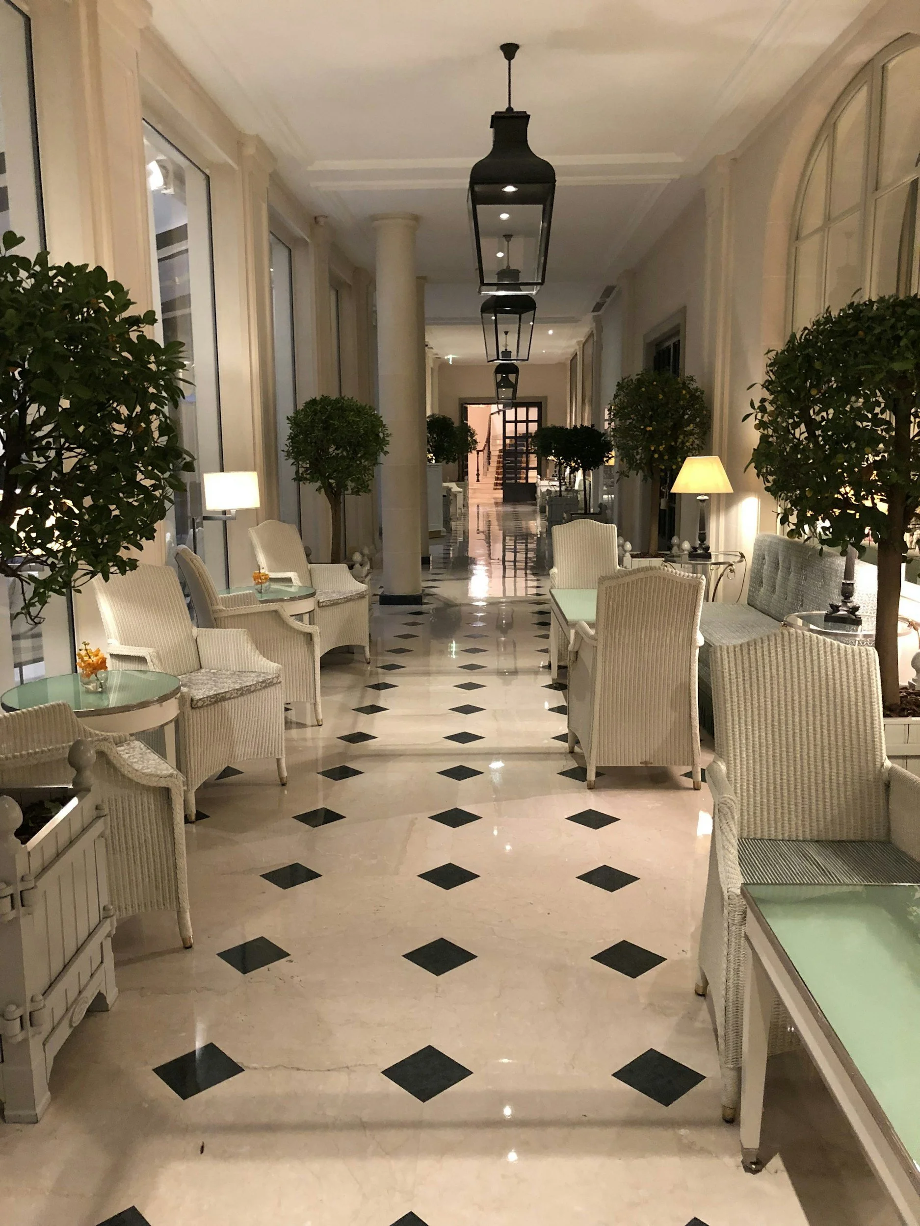 Hotel lobby with cream-colored chairs, green tables, potted plants, black and white checkered marble floor, and large windows with cream curtains. Hanging lanterns and lamps provide lighting.